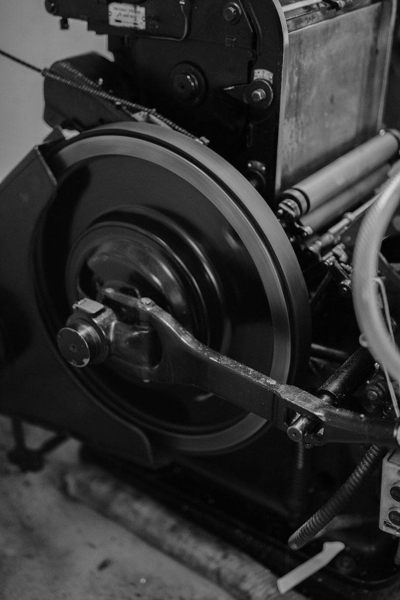 May include: A close-up of a large, black metal gear with a smaller gear attached to it. The gears are part of a printing press.