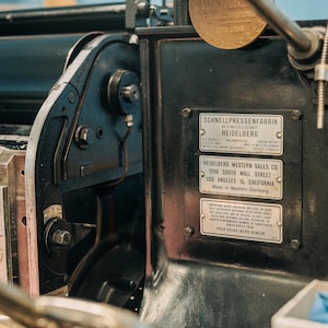 May include: Close-up of a vintage printing press with a black metal frame and a brass plate. The press has a label that reads "Schnellpressenfabrik Antengeseilschaft Heidelberg" and "Heidelberg Western Sales Co. 1700 South Wall Street Los Angeles 15, California Made in Western Germany".