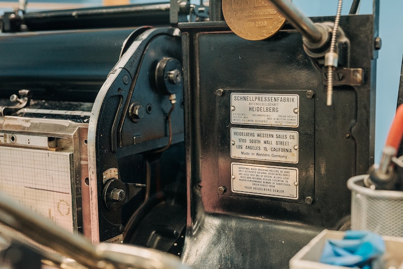 May include: Close-up of a vintage printing press with a black metal frame and a brass plate. The press has a label that reads "Schnellpressenfabrik Antengeseilschaft Heidelberg" and "Heidelberg Western Sales Co. 1700 South Wall Street Los Angeles 15, California Made in Western Germany".