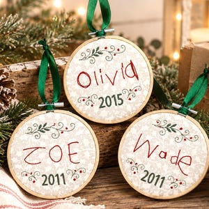 May include: Three circular wooden embroidery hoop ornaments featuring a light beige fabric with a subtle white floral pattern. Each ornament is embroidered with a name in red thread and a year in green thread, accented by green leaves and red berries.