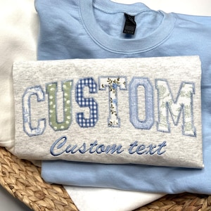 May include: A light blue sweatshirt featuring a heather gray t-shirt layered on top. The t-shirt displays the word CUSTOM in large, patterned fabric applique letters, with the phrase Custom text embroidered in blue script underneath.
