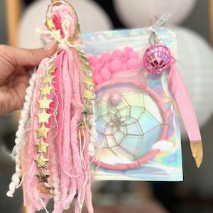 Peut inclure: Un ensemble attrape-rêves rose avec un pompon et un charme boule disco. L'attrape-rêves a un motif de toile d'araignée dorée et un cadre rose. Le pompon est rose et blanc avec des accents d'étoiles dorées. L'ensemble est emballé dans un sac irisé transparent.