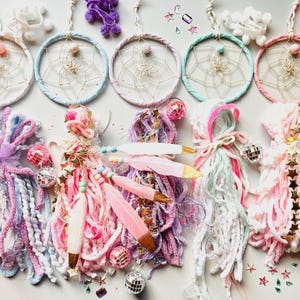 May include: A collection of handmade dream catchers in pastel colors. Each dream catcher features a circular hoop with intricate web designs, adorned with feathers, beads, and disco ball accents. The color palette includes pink, blue, and purple.