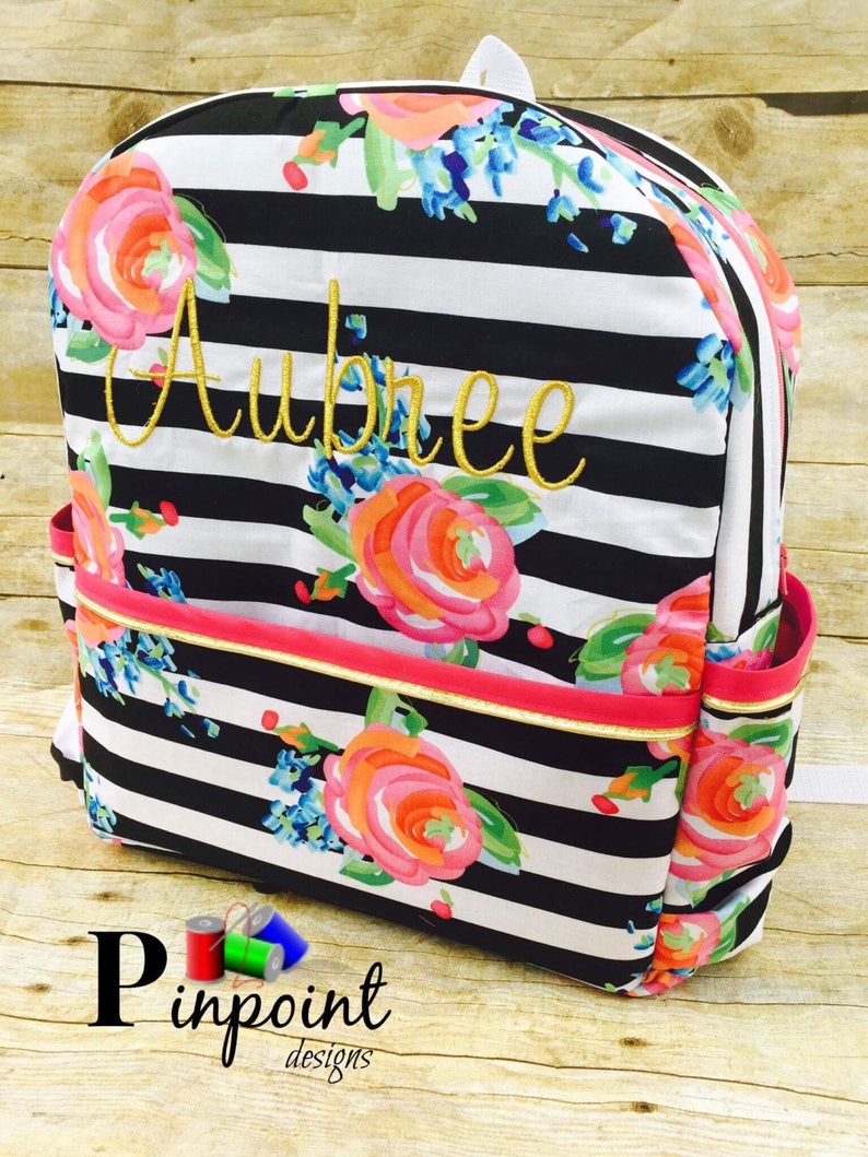 May include: Black and white striped backpack with a floral pattern and the name "Aubree" embroidered in gold thread. The backpack has a pink zipper and a gold trim.