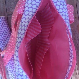 Puede incluir: Bolso de mano de tela rosa y morado con cierre de cremallera y varios bolsillos interiores. El bolso est&aacute; forrado con una tela rosa con rayas blancas.