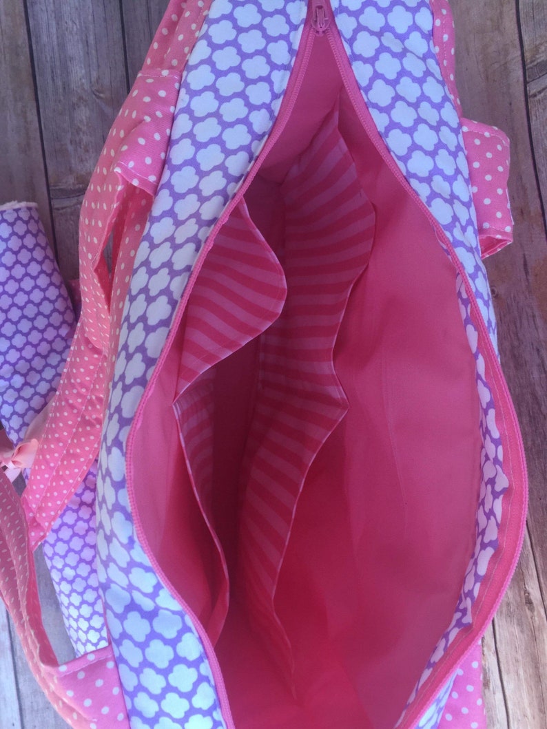 Puede incluir: Bolso de mano de tela rosa y morado con cierre de cremallera y varios bolsillos interiores. El bolso est&aacute; forrado con una tela rosa con rayas blancas.