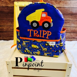May include: Blue and yellow construction vehicle backpack with the name "TRIPP" embroidered on the front. The backpack has a pocket on the front and a small pocket on the side. The backpack is sitting on a wooden crate.
