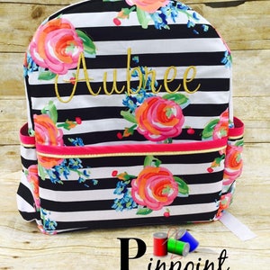 May include: Black and white striped backpack with a floral pattern and the name "Aubree" embroidered in gold thread. The backpack has a pink zipper and a gold trim.