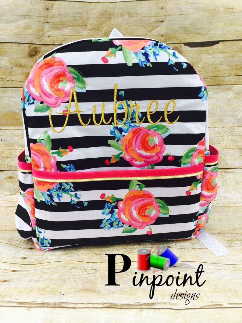 May include: Black and white striped backpack with a floral pattern and the name "Aubree" embroidered in gold thread. The backpack has a pink zipper and a gold trim.