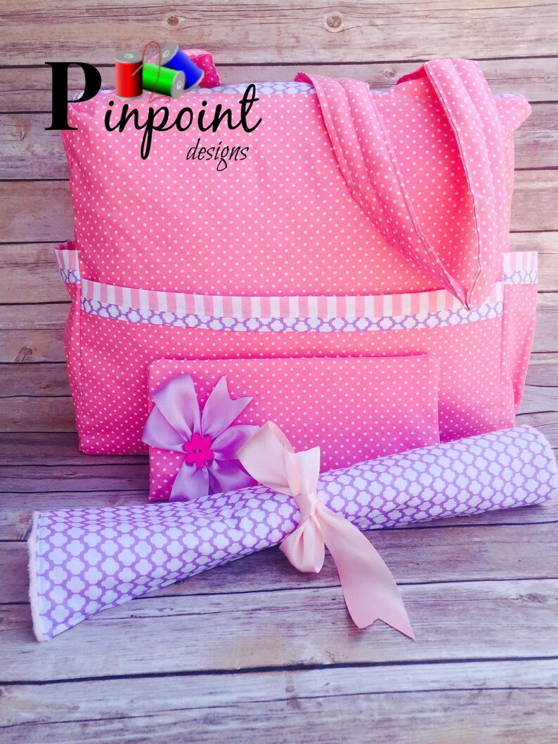 Puede incluir: Bolso tote de lunares rosa con ribete a rayas morado y blanco y una bolsa m&aacute;s peque&ntilde;a a juego. El bolso tote tiene un asa de tela rosa y la bolsa tiene una cinta rosa atada alrededor. El bolso tote tiene el texto "Pinpoint Designs" en la parte delantera.