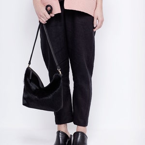 Puede incluir: Una mujer lleva un c&aacute;rdigan rosa, pantalones negros y mules negros de tac&oacute;n. Lleva un bolso de mano de piel negra con una correa larga.