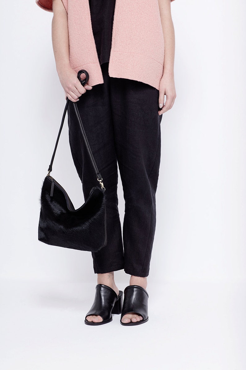Puede incluir: Una mujer lleva un c&aacute;rdigan rosa, pantalones negros y mules negros de tac&oacute;n. Lleva un bolso de mano de piel negra con una correa larga.
