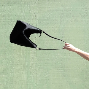 Puede incluir: Un bolso de mano de cuero negro con una correa larga, colgando de una mano contra una pared verde.