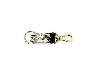 May include: A black and white cowhide leather keychain with a gold metal key ring and a gold metal clip.