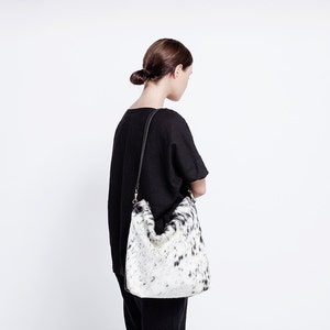 Puede incluir: Una mujer con una camisa negra y pantalones negros lleva un bolso tote de piel de vaca blanca y negra con una correa negra.