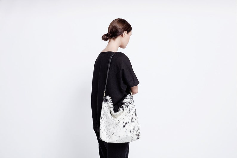 Puede incluir: Una mujer con una camisa negra y pantalones negros lleva un bolso tote de piel de vaca blanca y negra con una correa negra.