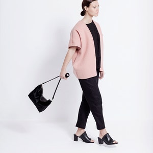 Puede incluir: Una mujer con un c&aacute;rdigan rosa, pantalones negros y mules negras de tac&oacute;n. Lleva un bolso negro con una correa larga.