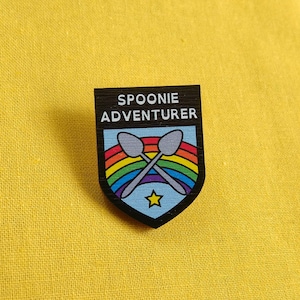 May include: Black and white pin with a rainbow design and the text "Spoonie Adventurer" with two crossed spoons in the centre.