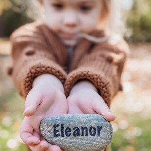 Peut inclure: Les mains d'un enfant tenant une pierre grise avec le nom "Eleanor" &eacute;crit en lettres noires.