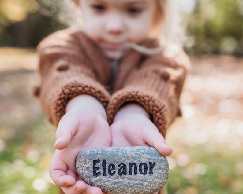 Peut inclure: Les mains d'un enfant tenant une pierre grise avec le nom "Eleanor" &eacute;crit en lettres noires.