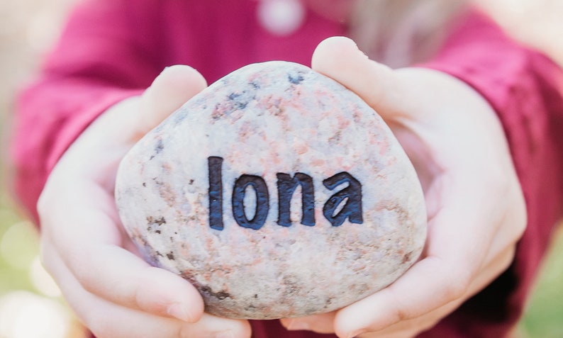 Peut inclure: Gros plan d'une main tenant une pierre grise avec le nom "Iona" grav&eacute; en lettres noires.