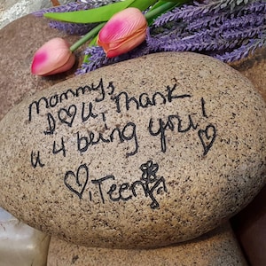 以下が含まれることがあります： 黒いインクで「Mommy I love you Thank you 4 being you」と書かれた、茶色の楕円形の石。石には小さな花とハートも描かれている。