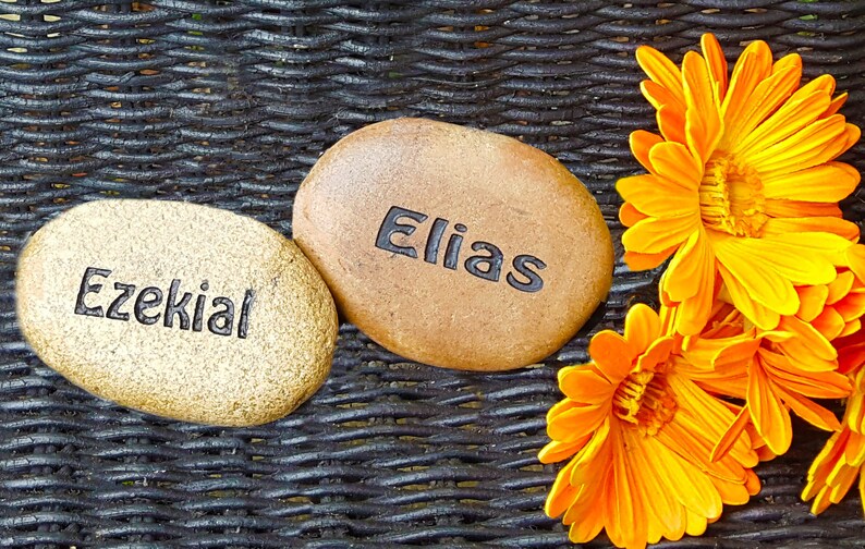 Peut inclure: Deux pierres lisses de forme ovale avec les noms "Ezekial" et "Elias" grav&eacute;s dessus. Les pierres reposent sur une surface en osier tress&eacute;, entour&eacute;es de fleurs orange.