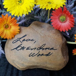以下が含まれることがあります： 「Love, Breanna Wood」と刻まれた茶色の石。石は黄色、オレンジ、赤の花に囲まれている。
