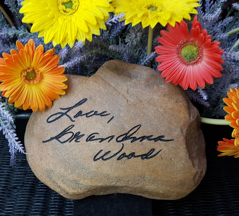 以下が含まれることがあります： 「Love, Breanna Wood」と刻まれた茶色の石。石は黄色、オレンジ、赤の花に囲まれている。