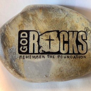 以下が含まれることがあります： 灰色と茶色の石に、「GOD ROCKS」と「REMEMBER THE FOUNDATION」という文字が黒で刻まれています。