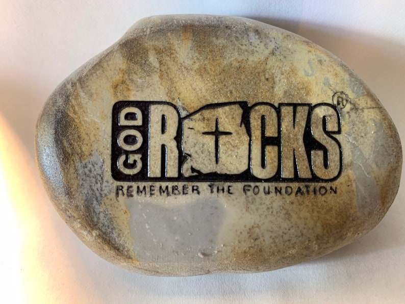 以下が含まれることがあります： 灰色と茶色の石に、「GOD ROCKS」と「REMEMBER THE FOUNDATION」という文字が黒で刻まれています。