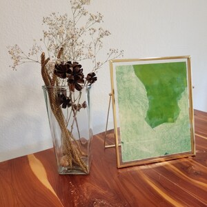 Könnte beinhalten: Ein goldfarbener Bilderrahmen mit einem grünen abstrakten Design steht auf einem Holztisch. Neben dem Bild steht eine Glasvase mit getrockneten Blumen und braunen Tannenzapfen.