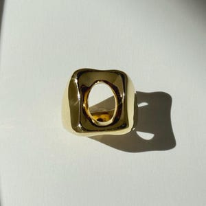 May include: A gold ring with a square design and a rounded, open centre. The ring has a smooth, polished surface and casts a shadow on a white surface. The ring is a piece of jewellery.
