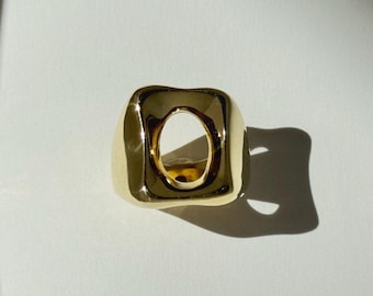 May include: A polished gold-tone ring featuring a thick, rounded square band with a prominent, smooth, oval-shaped cutout in the center. The metal surface is highly reflective, creating bright highlights and deep shadows on its curved form.