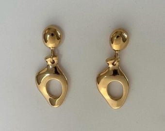 May include: A pair of gold-colored earrings. Each earring features a small, round stud connected to a teardrop-shaped pendant with a circular cutout. The earrings have a polished, reflective surface.