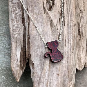 May include: A silver necklace with a cat pendant. The pendant is a dark red color with black speckles. The cat is in a sitting position with its tail curled. The necklace is displayed on a piece of weathered wood.