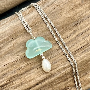 May include: A silver necklace with a cloud-shaped pendant in light blue and a white teardrop-shaped pearl. The necklace is displayed on a wooden surface.