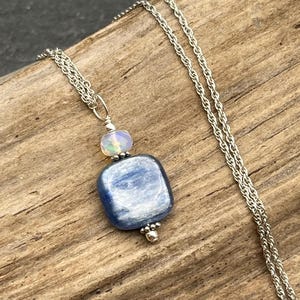 May include: A silver chain necklace featuring a pendant with a square, polished blue kyanite stone and a smaller, translucent, iridescent opal bead. The pendant is accented with small silver beads at the top and bottom, suspended from a delicate silver chain.