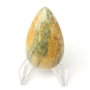 May include: A teardrop-shaped stone with a mix of yellow, brown, and green hues. The polished stone is displayed on a clear acrylic stand, showcasing its natural colors and textures. The stone's shape is smooth and elongated, with a rounded top.