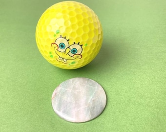 May include: A yellow golf ball with a cartoon face and a round, light-colored stone. The golf ball has a dimpled surface and the cartoon face is smiling. The stone is circular and has a marbled appearance.