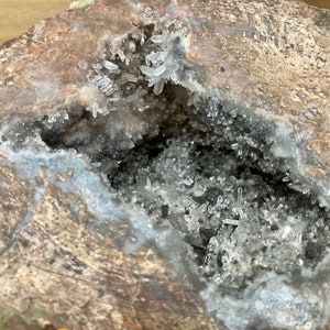 Puede incluir: Una geoda con una gran abertura que revela un cúmulo de cristales de cuarzo blanco en su interior. La geoda es marrón y gris con una superficie rugosa y texturizada.
