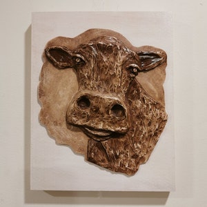 May include: A brown cow's head sculpture mounted on a white wooden plaque. The sculpture is made of a textured material that resembles wood or bark.
