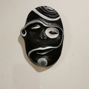 May include: Black and white ceramic mask with a stylised face and abstract patterns. The mask has a closed eye and a white circle around the other eye.