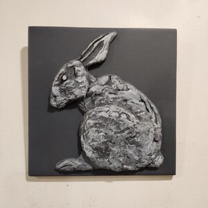 May include: A grey rabbit sculpture with a textured surface, mounted on a black square background.