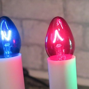 May include: Four Christmas light bulbs with different colored glass and a letter inside each bulb. The bulbs are blue, red, green, and red. The letters inside the bulbs are N, A, N, and M.