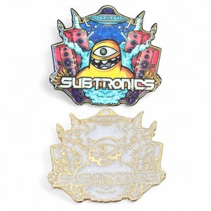 May include: Two enamel pins featuring a cartoon alien design. The top pin is colorful with a yellow alien, speakers, and the word "SUBTRONICS". The bottom pin is gold and white with the same design.