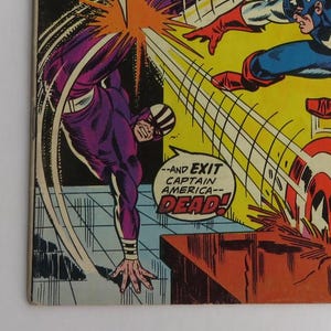 Puede incluir: Un panel de c&oacute;mic que muestra a un personaje de superh&eacute;roe morado y negro pateando a un personaje de superh&eacute;roe rojo, blanco y azul. El texto en el panel dice "--and exit captain america-- dead!"