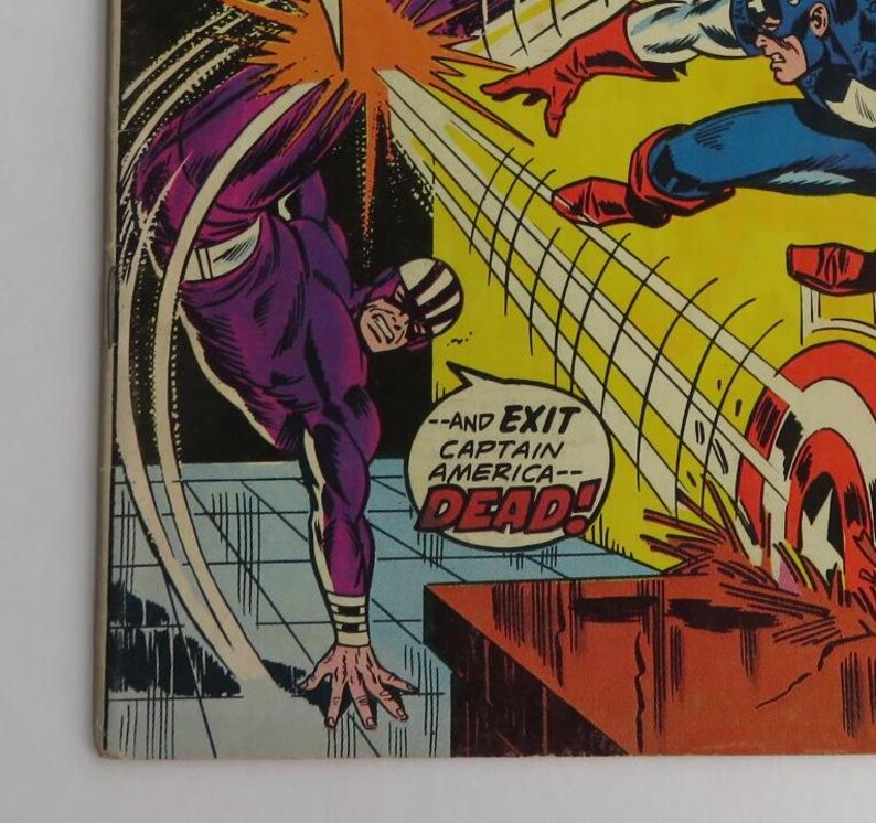 Puede incluir: Un panel de c&oacute;mic que muestra a un personaje de superh&eacute;roe morado y negro pateando a un personaje de superh&eacute;roe rojo, blanco y azul. El texto en el panel dice "--and exit captain america-- dead!"