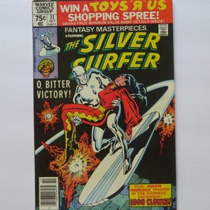May include: A comic book cover featuring the Silver Surfer, a silver humanoid character, riding a surfboard through space. The Surfer is carrying a woman with long black hair, who is wearing a red and yellow costume. The cover text reads "The Silver Surfer" and "O, Bitter Victory!"