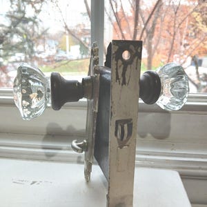 May include: An antique door lock set with clear glass doorknobs and a metal backplate. The lock has a tarnished, aged appearance, with visible wear and tear. The doorknobs have a faceted design.